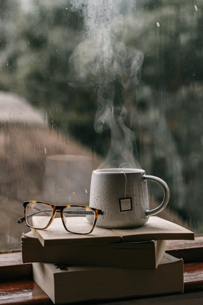 Comforting setup with books, glasses, and hot tea by a rain-drenched window, perfect for reading.